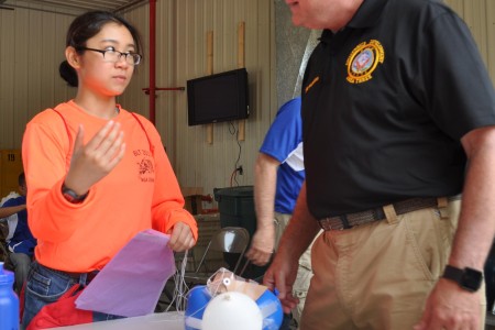 A Navy Junior ROTC student attending the Basic Leadership Training summer camp explains her team's egg drop project. Cadets worked together to create a contraption that would protect the egg during a drop from a three story building without breaking or cracking the egg after learning about the Mars Spirit Rover Landing. The BLT camp was held at Crane Army Ammunition Activity's reserve facility located on Naval Support Activity Crane June 26-30. 